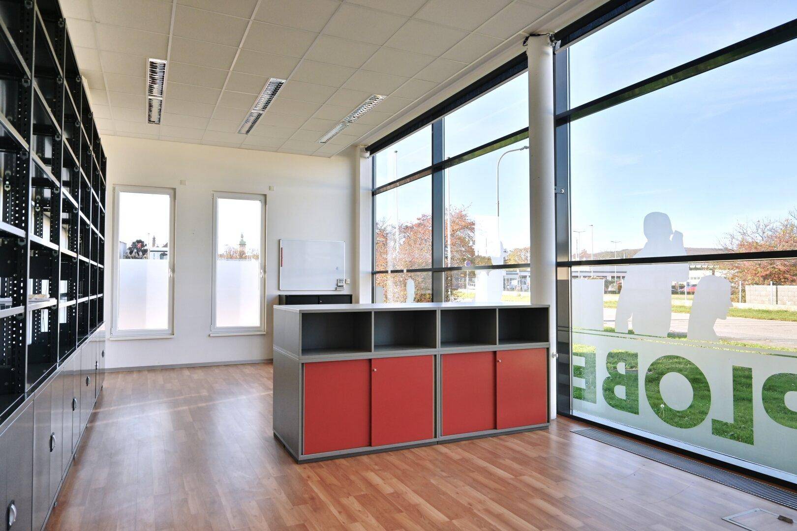 Heller Büroraum mit großen Fenstern, Holzfußboden und modernen roten Schränken, die viel Stauraum bieten.