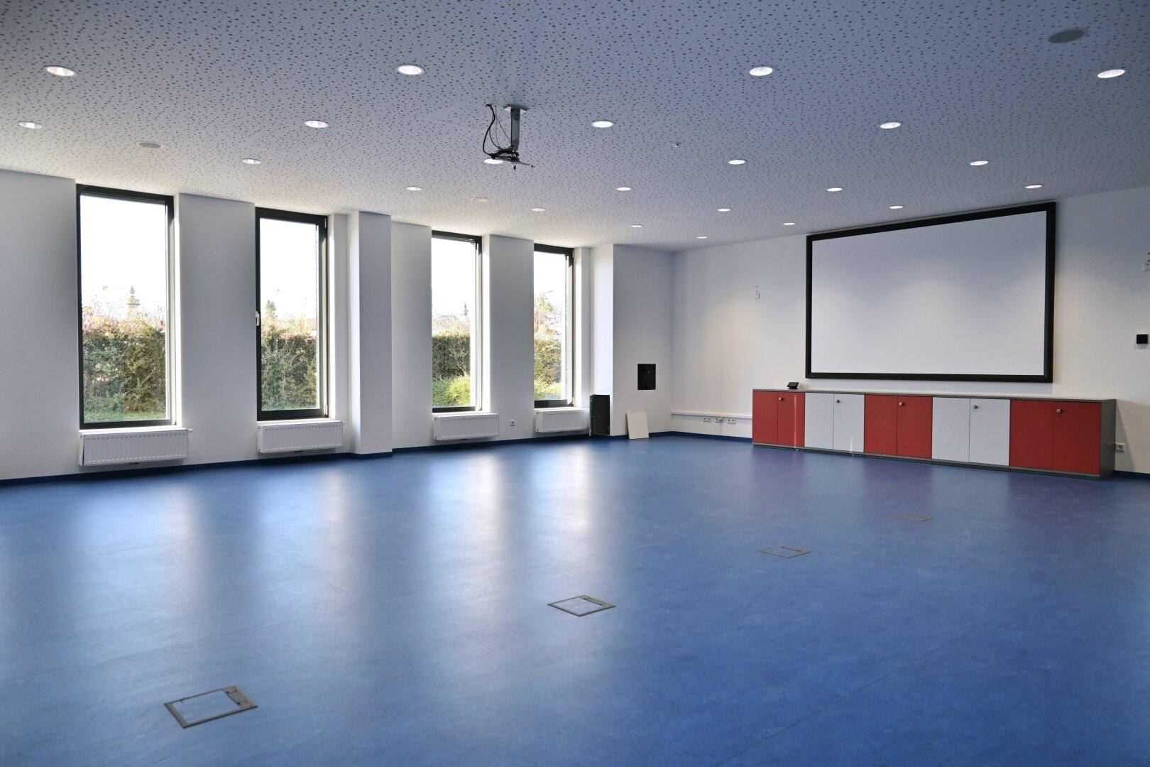 Großer Konferenzraum mit blauem Boden, Projektionsfläche und hohen Fenstern für viel Tageslicht.