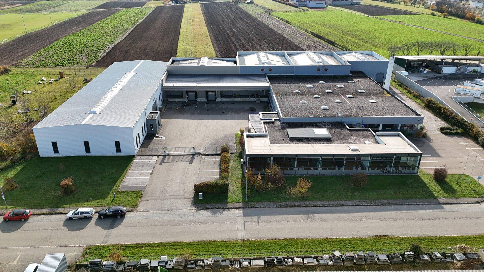Weitläufiger Gewerbekomplex mit Parkplätzen und angrenzenden landwirtschaftlichen Flächen unter blauem Himmel.