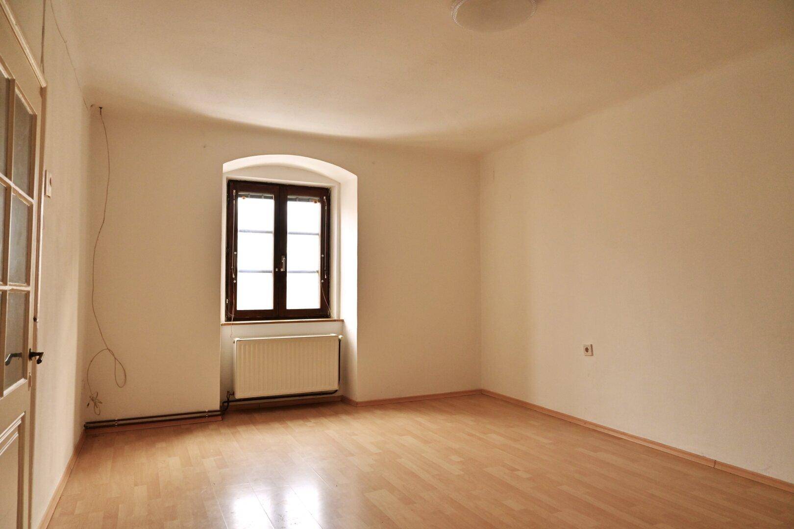 Leeres Zimmer mit Holzboden, einem Fenster und einem Heizkörper, bereit zur individuellen Gestaltung.
