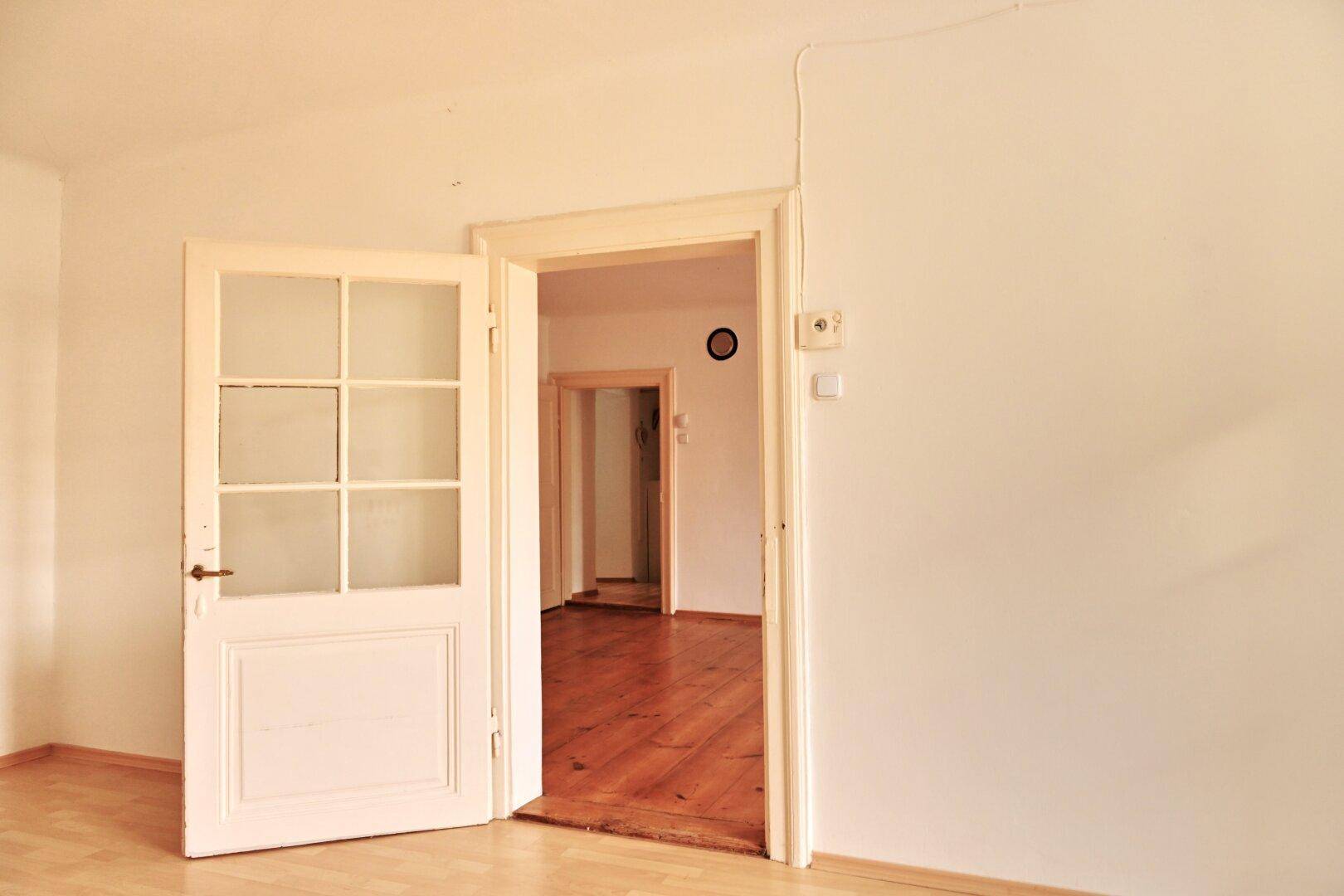 Raum mit Holzboden und offener Tür, die den Blick in einen weiteren Bereich der Wohnung freigibt.