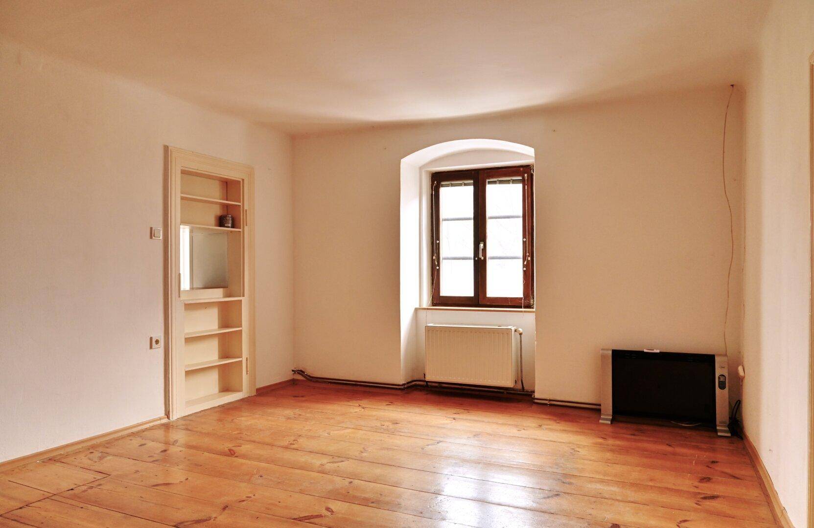 Zimmer mit Holzboden, Fenster, Heizkörper und einem praktischen Einbauregal für Stauraum.