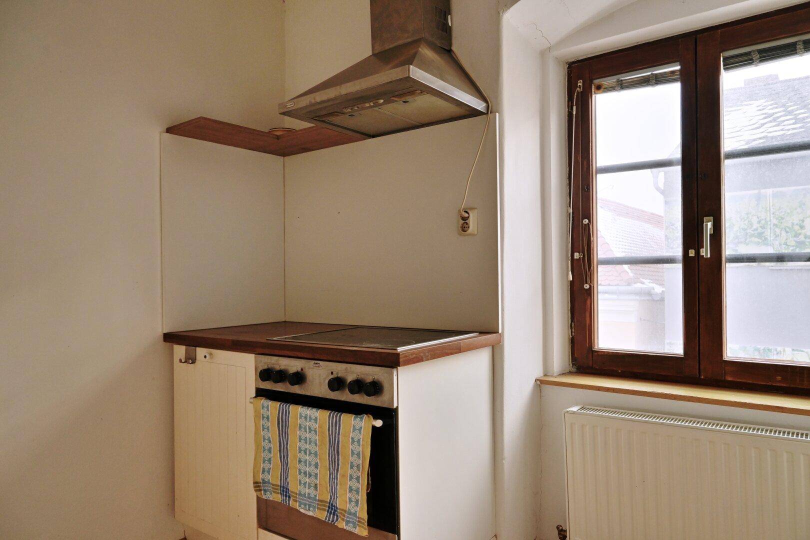 Küchenbereich mit Herd, Backofen, Dunstabzugshaube und einem Fenster, das Tageslicht spendet.