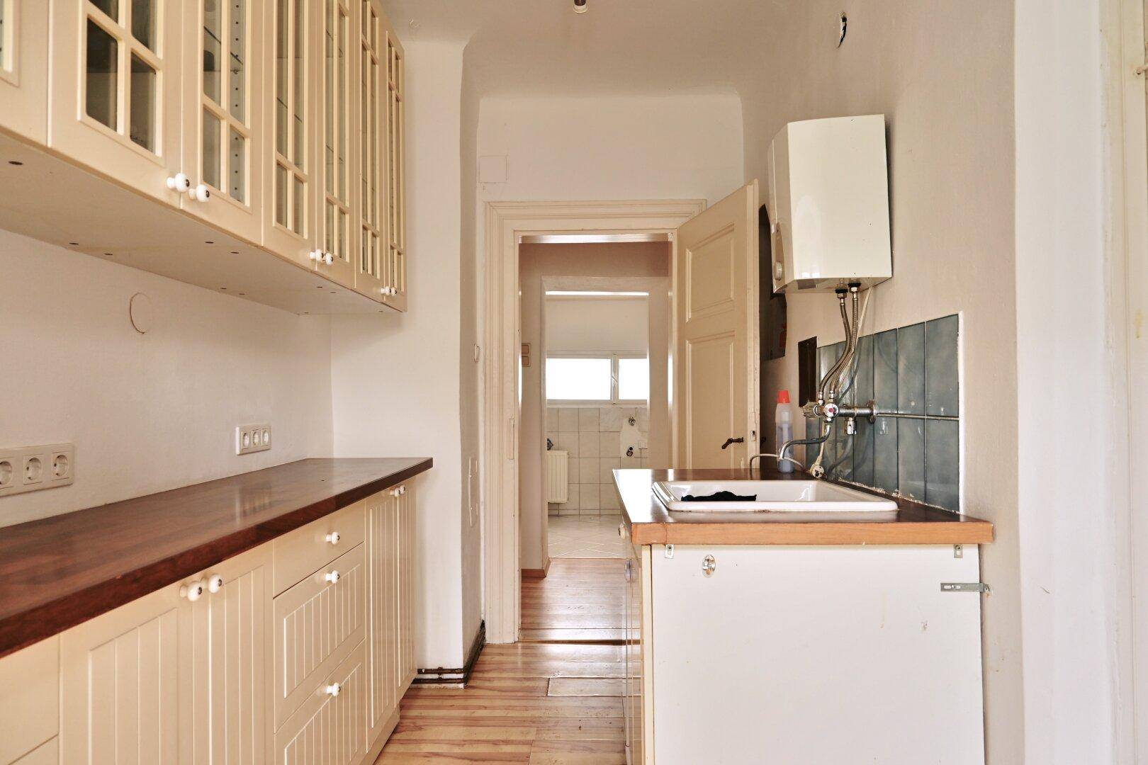 Schmale Küche mit hellen Holzschränken, Holzarbeitsplatte und Blick in einen angrenzenden Raum.