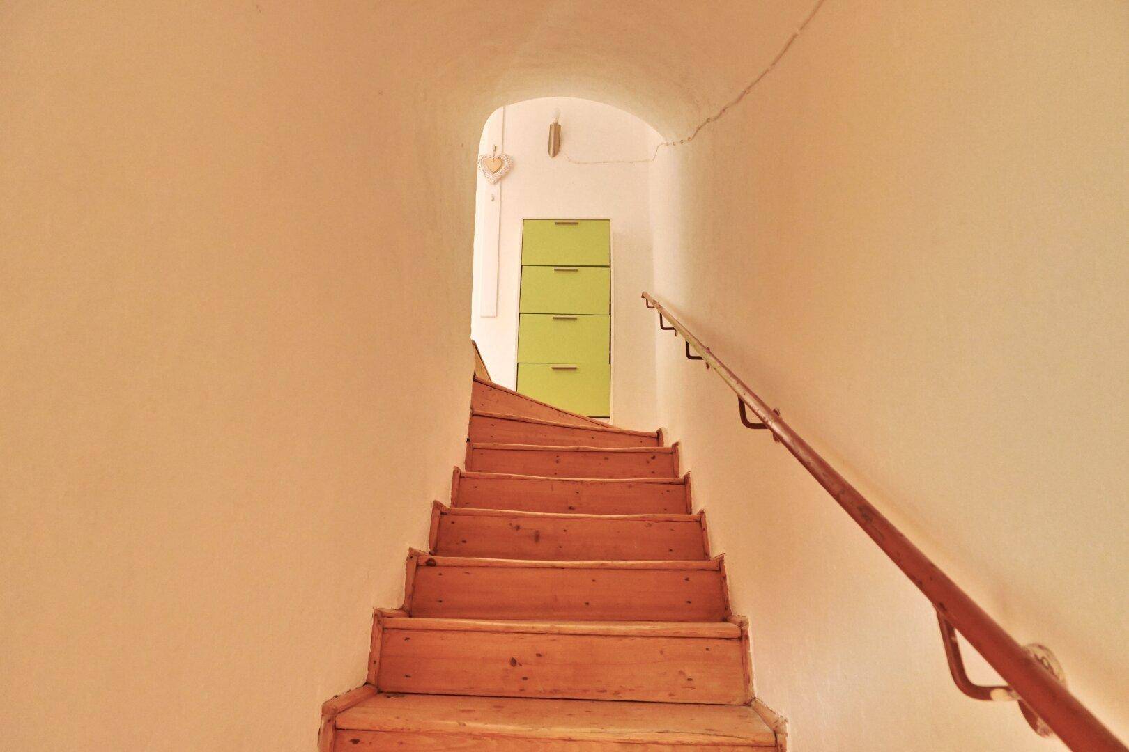 Holztreppe mit Geländer führt nach oben, am Ende ist ein grüner Schrank sichtbar.