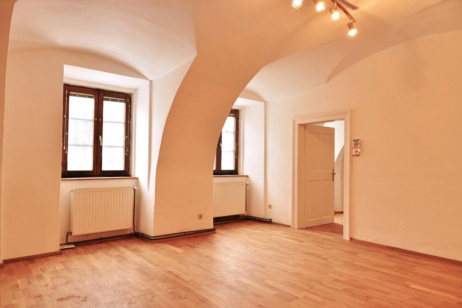 Heller Wohnraum mit Holzboden, Gewölbedecken und zwei Fenstern, die viel Tageslicht hereinlassen.