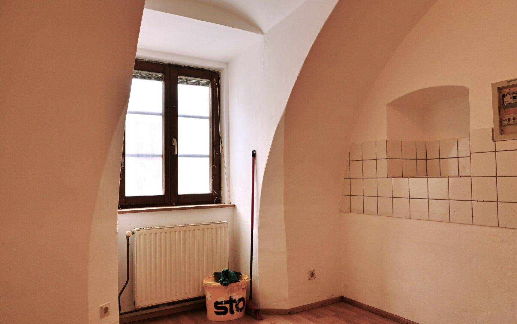 Ecke eines Raumes mit Fenster, Heizkörper und teilweise gefliesten Wänden unter einer Gewölbedecke.