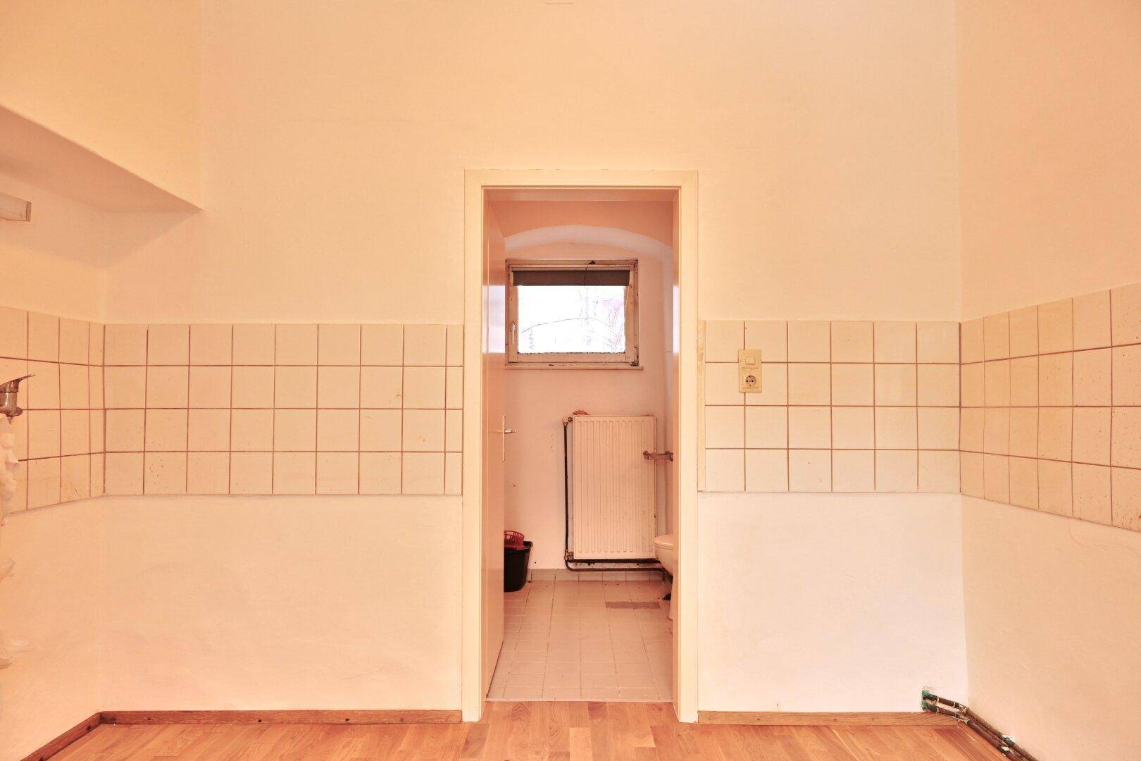 Durchgang zu einem kleinen Raum mit Fenster, umgeben von teilweise gefliesten Wänden im Vordergrund.