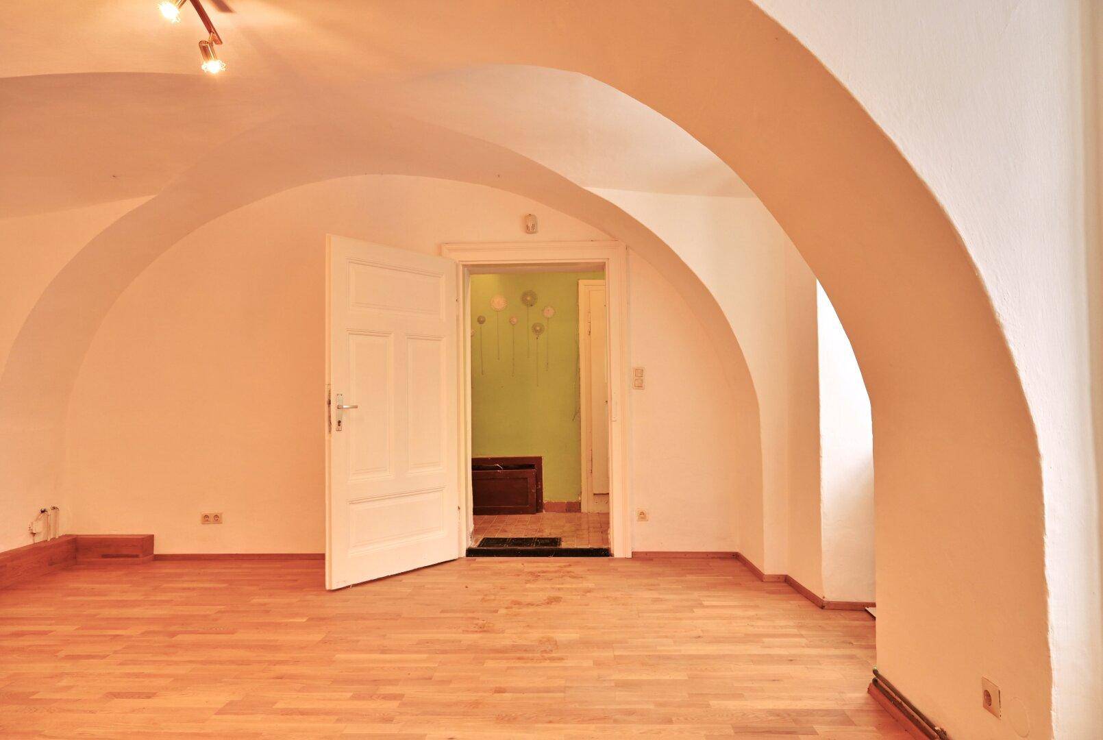 Geräumiger Raum mit Holzboden und Gewölbedecke, eine offene Tür führt zu einem grün gestrichenen Zimmer.