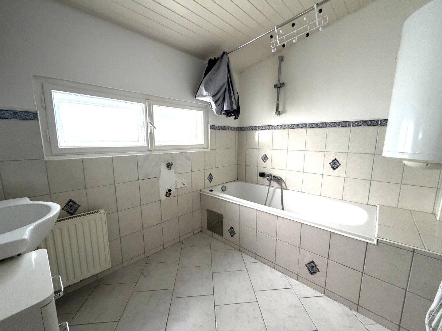 Helles Badezimmer mit Badewanne, Waschbecken, Heizkörper und Fenster, gefliest in Weiß und Blau.