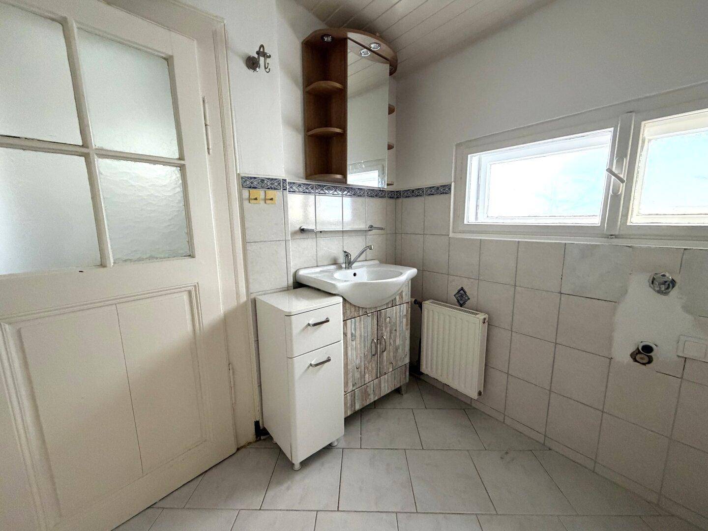 Badezimmer mit Waschbecken, Spiegelschrank, Heizkörper und Fenster, gefliest in hellen Farben.