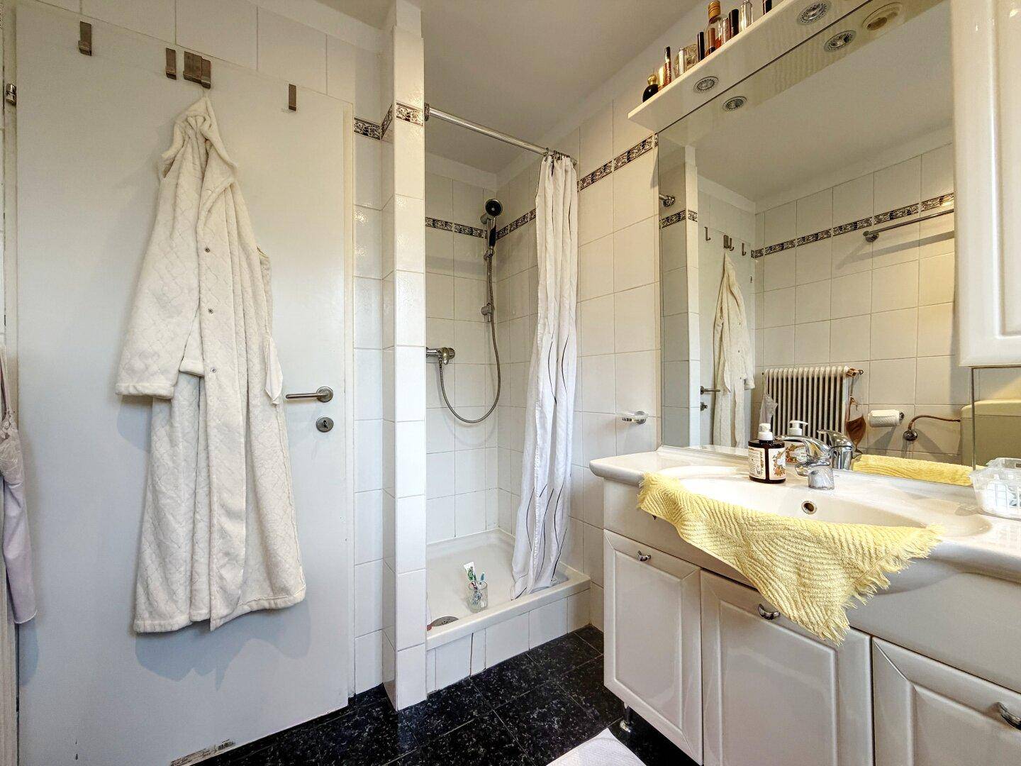 Badezimmer mit Dusche, Waschbecken und Spiegel, komplett weiß gefliest.