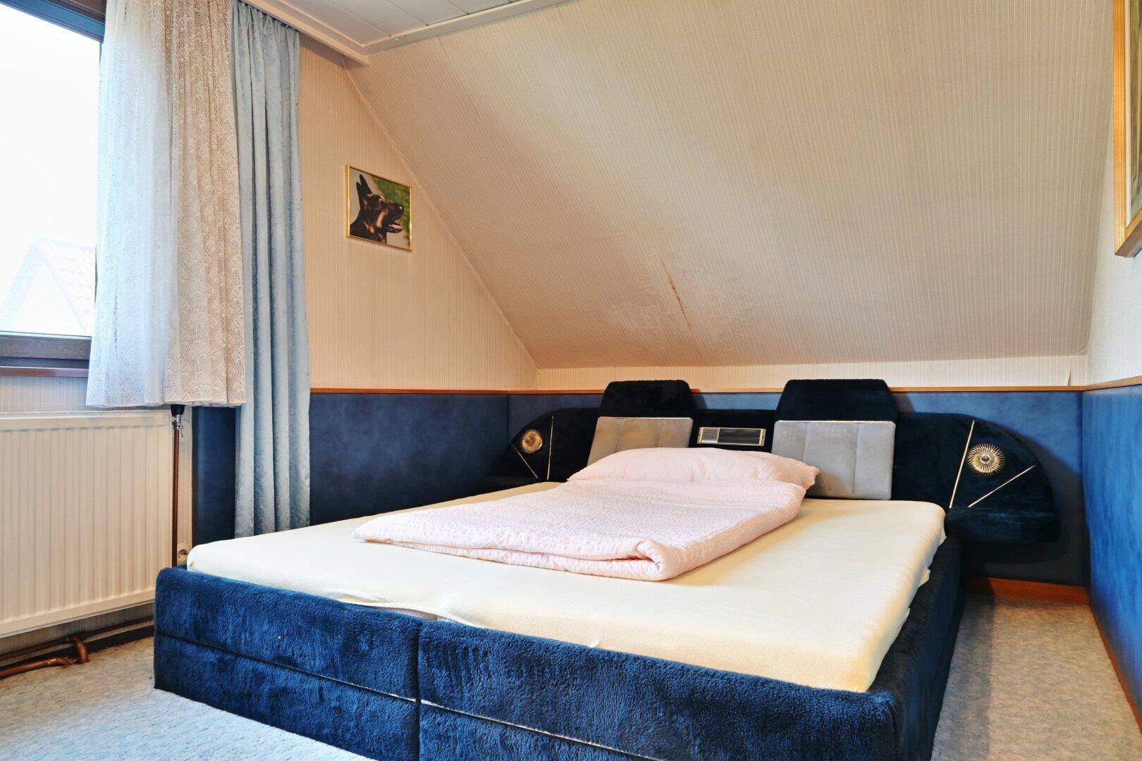 Schlafzimmer im Dachgeschoss mit blauem Bett, schrägen Wänden und Teppichboden.