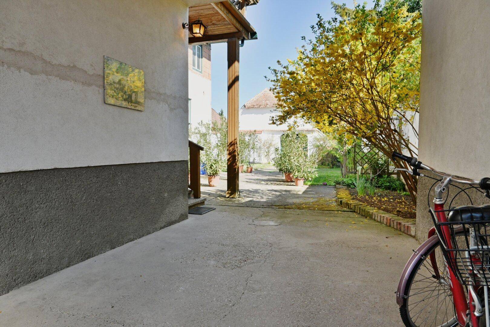 Gepflasterter Weg führt durch den Gartenbereich mit blühenden Sträuchern und Topfpflanzen.