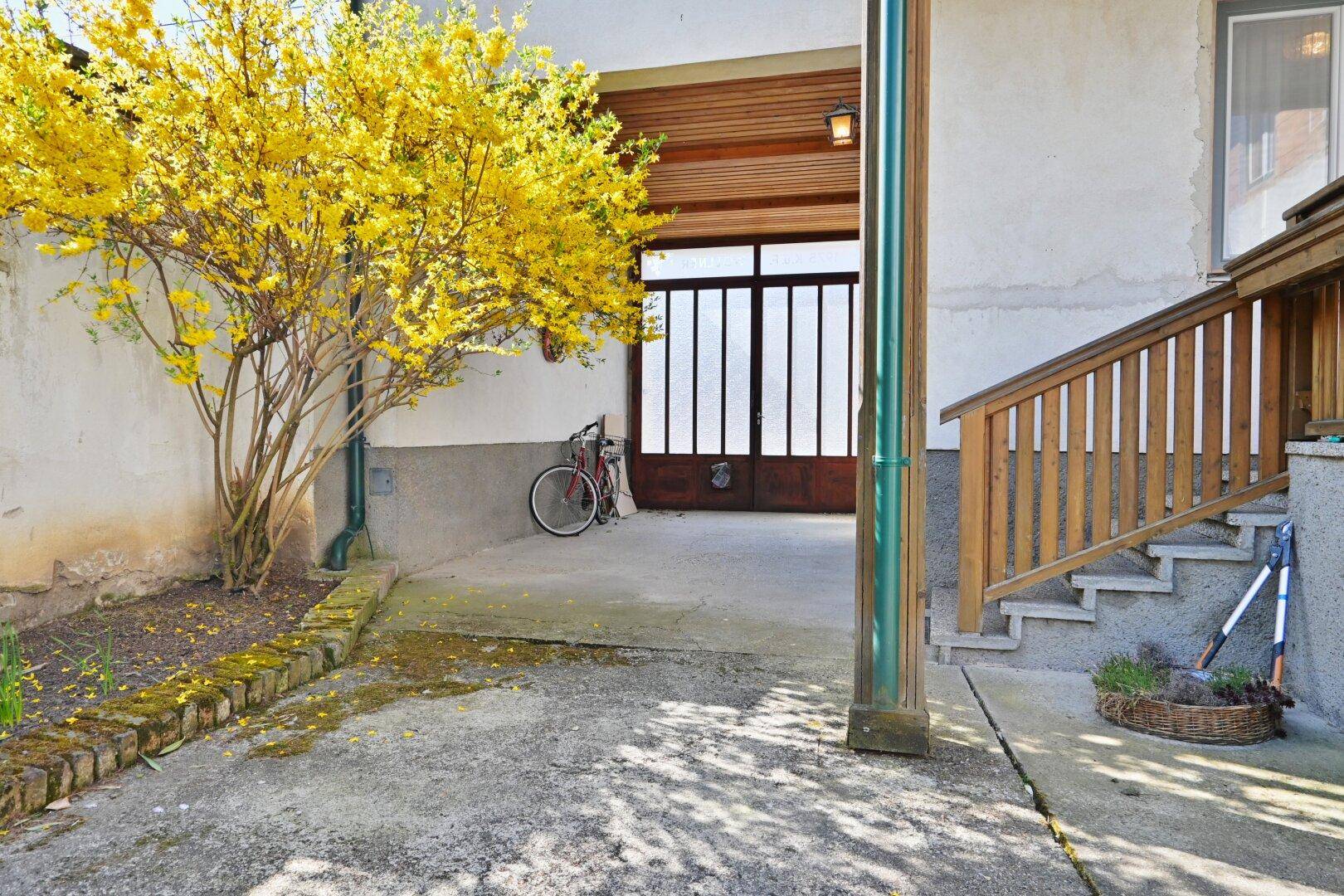 Eingangsbereich im Freien mit Holztreppe, einem blühenden Baum und Zugang zu einer Garage.