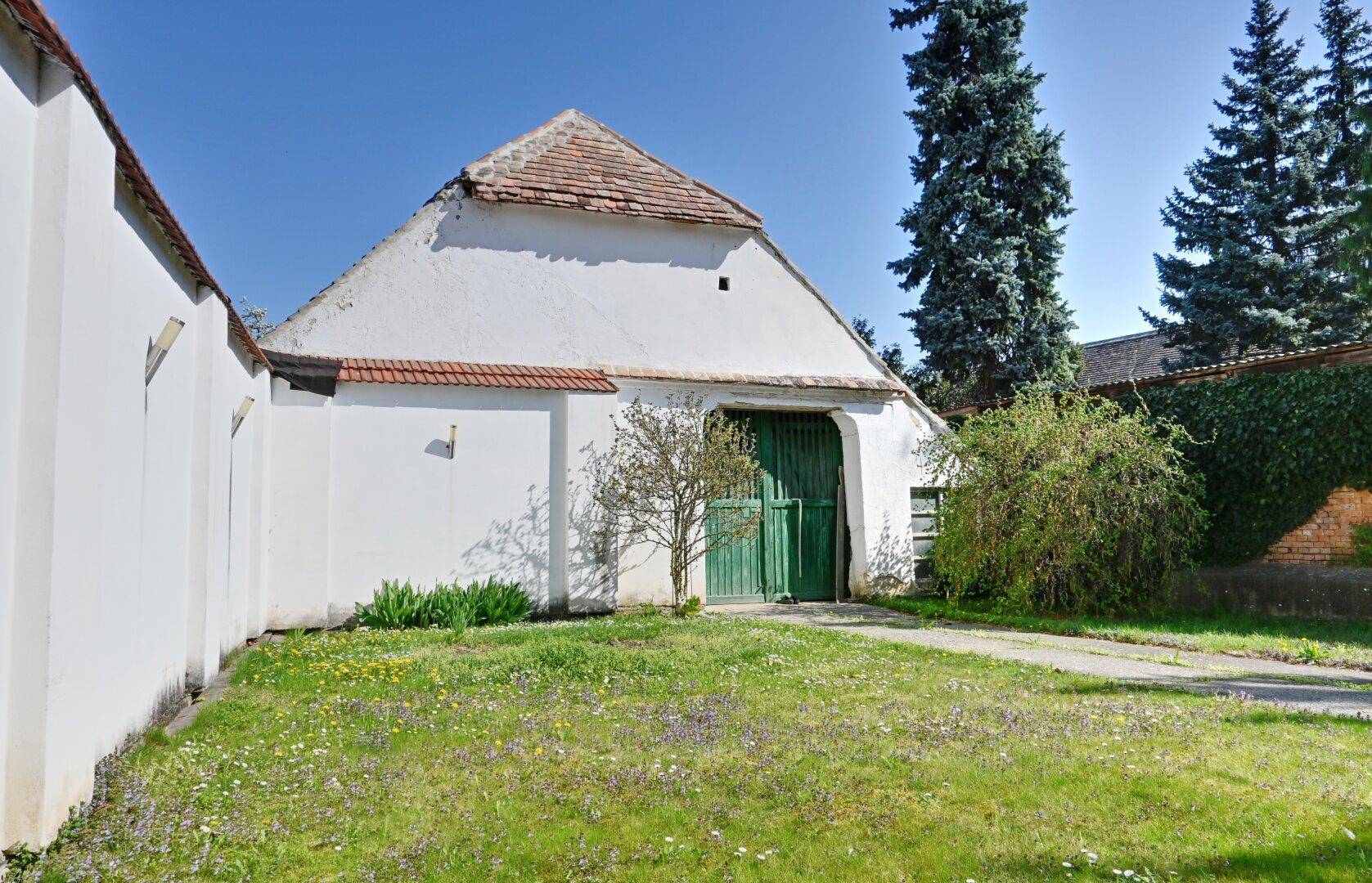 Weißes Nebengebäude mit grünen Holztoren und einer Rasenfläche im sonnigen Garten.