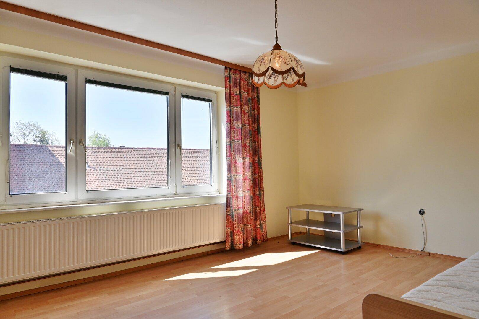 Helles Schlafzimmer mit Parkettboden, großem Fenster und Blick auf die Dächer.