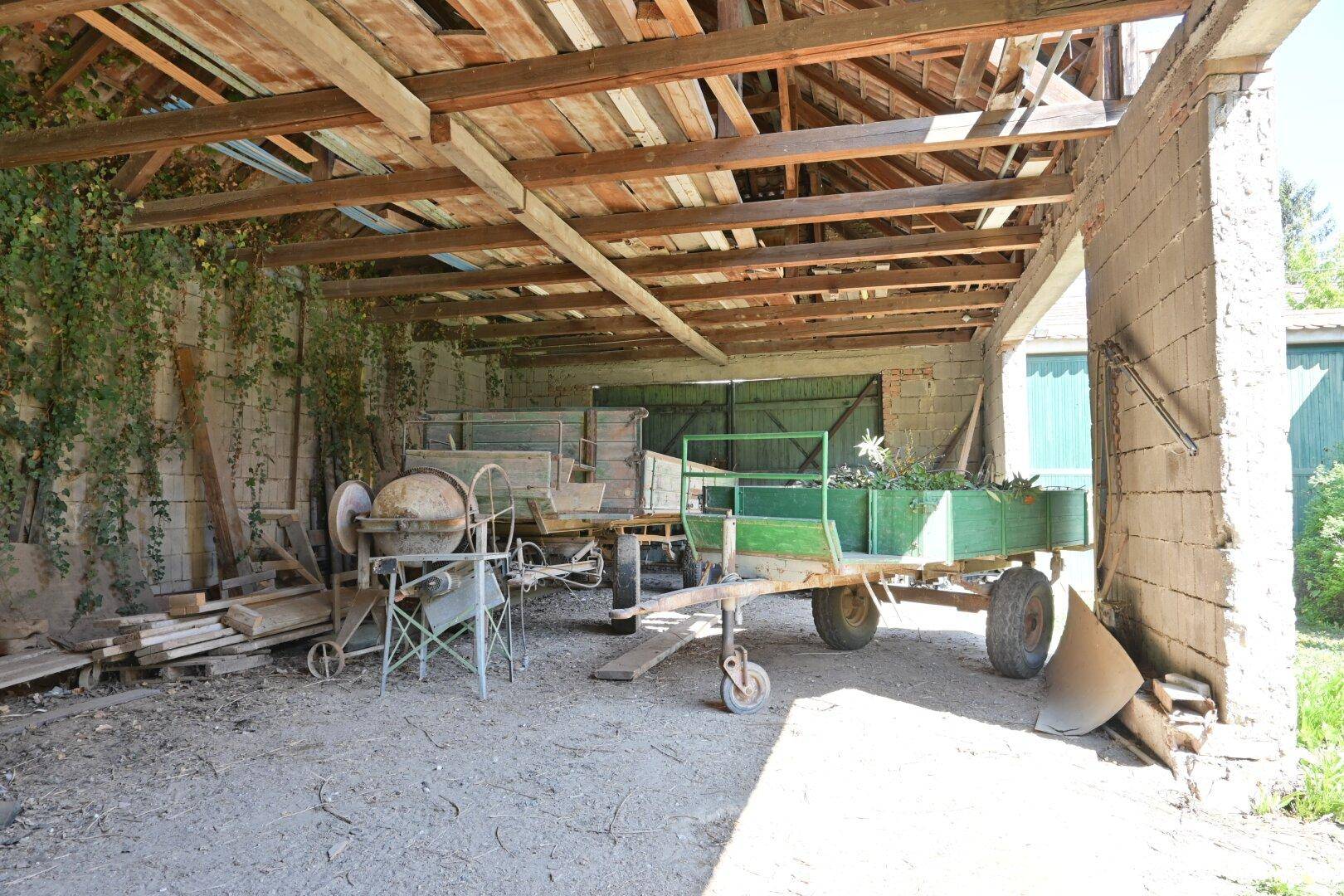Rustikaler Unterstand mit Holzbalken und landwirtschaftlichen Geräten, umgeben von Grün.