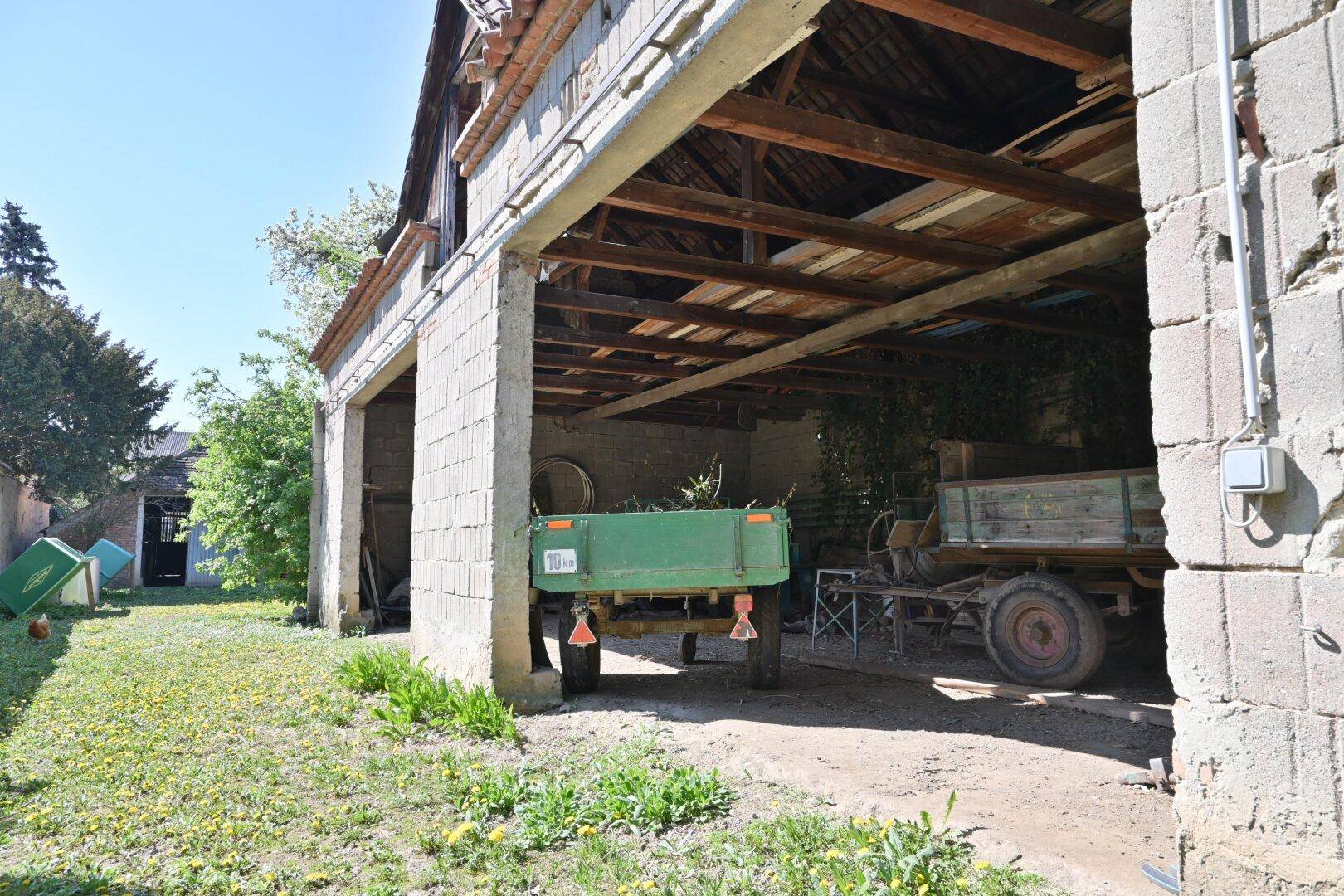 Großer Unterstand mit Holzdachkonstruktion und landwirtschaftlichem Anhänger auf dem Gelände.