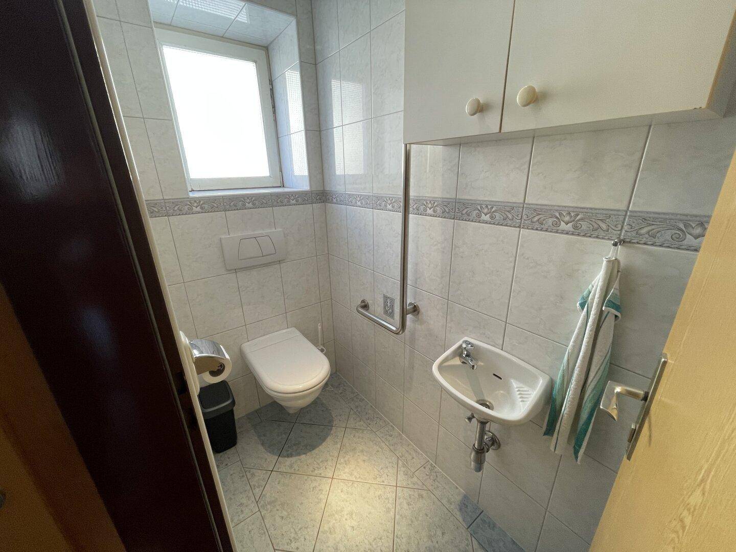 Kleines WC mit Fenster, Wand-WC, Handwaschbecken und Haltegriffen.