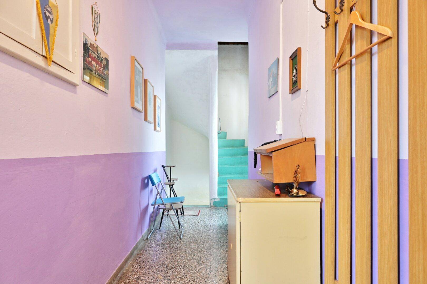 Flur mit lila und weiß gestrichenen Wänden, Terrazzoboden und einer Treppe im Hintergrund.