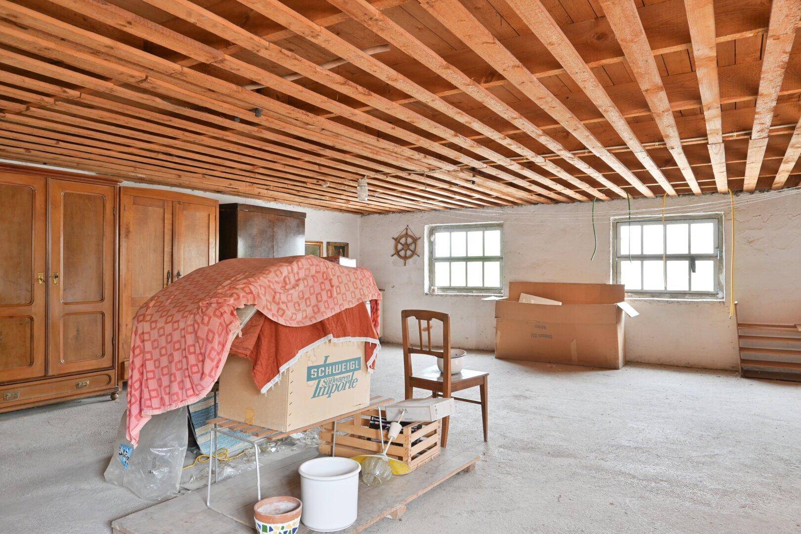 Dachbodenraum mit sichtbaren Holzdeckenbalken, Betonboden und Fenstern, die Licht spenden.
