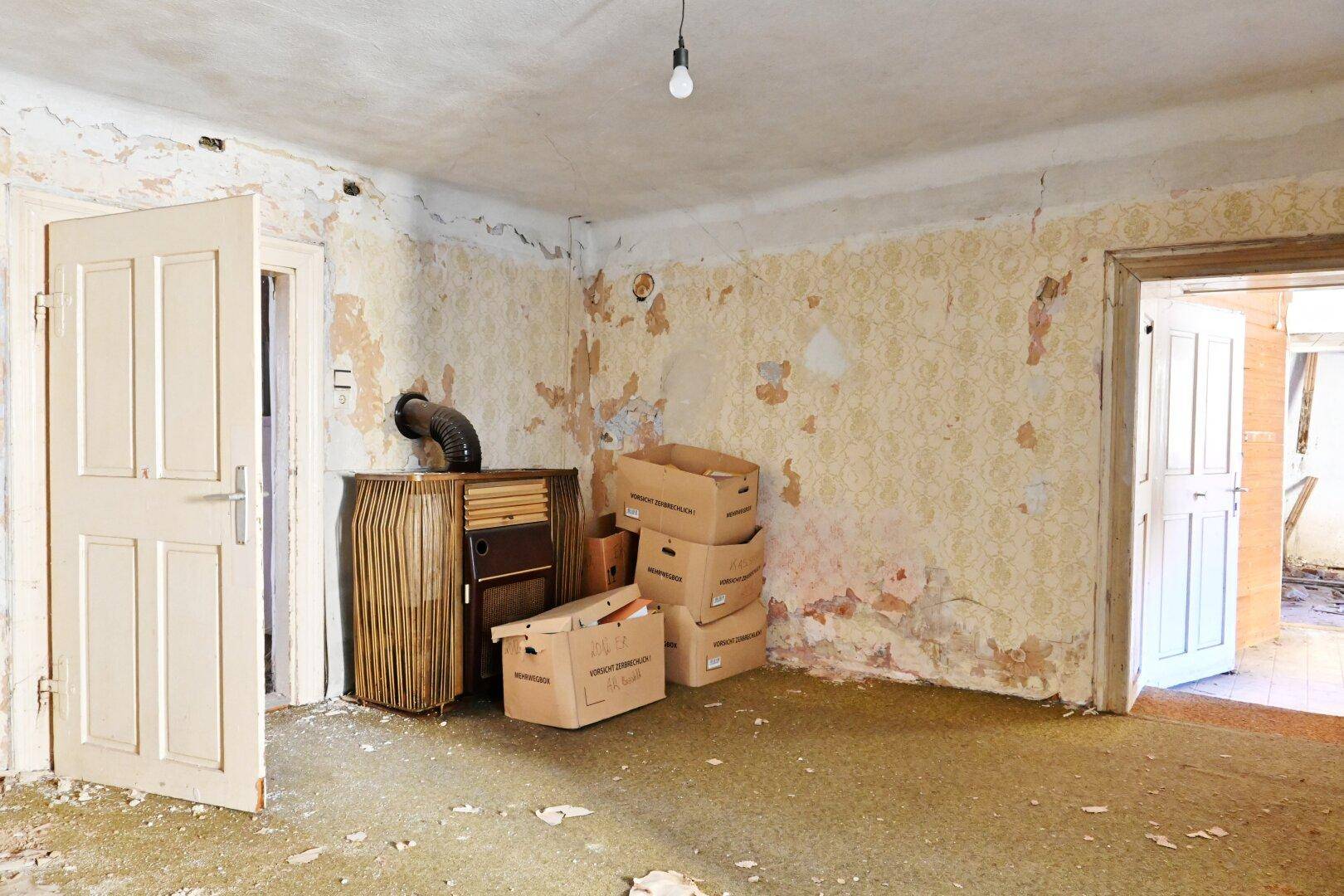 Leerer Raum mit abblätternder Tapete und alten Möbeln, der umfassende Renovierungsarbeiten erfordert.