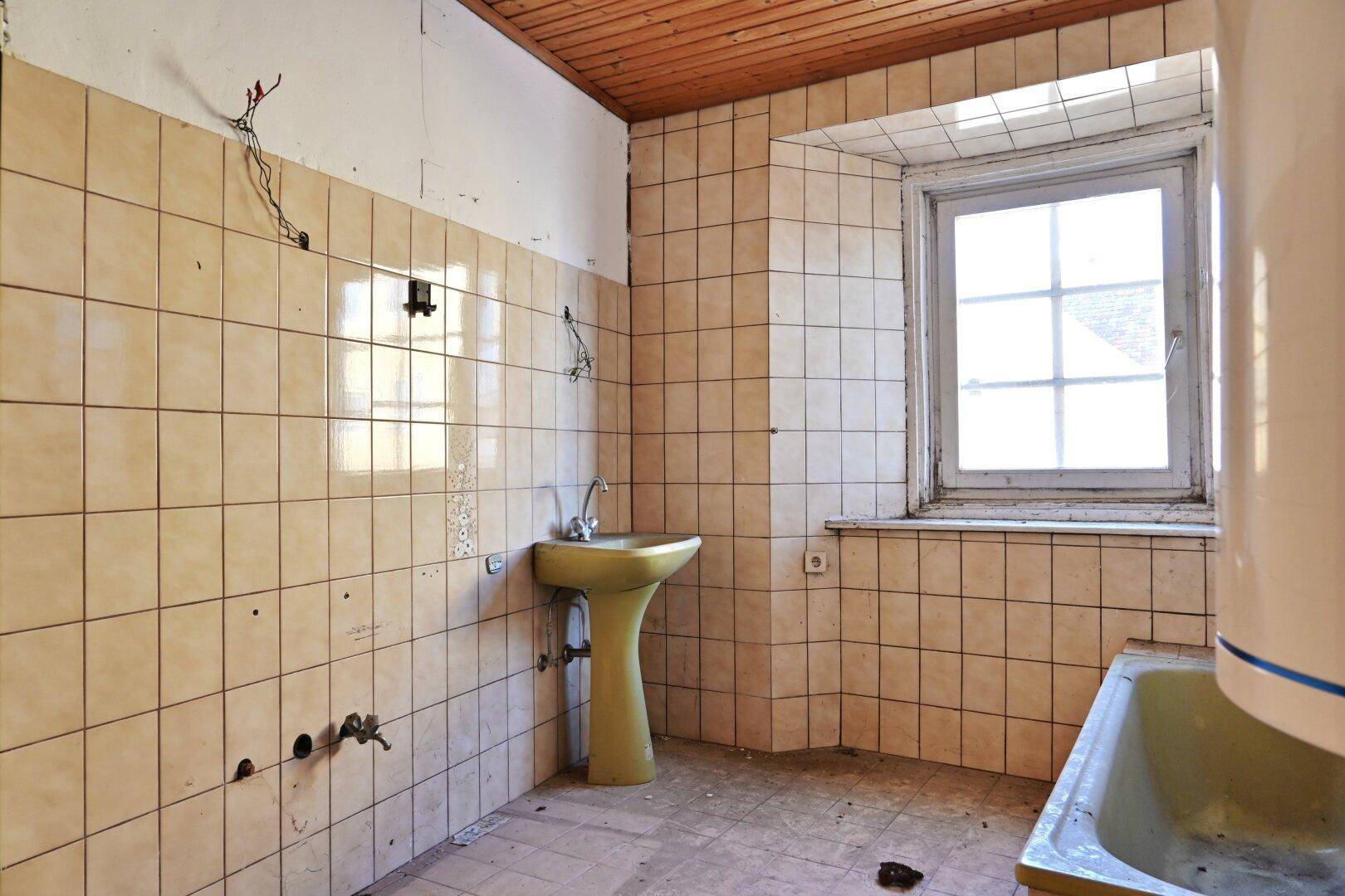 Stark sanierungsbedürftiges Badezimmer mit gelben Fliesen, Waschbecken und Badewanne sowie einem Fenster.