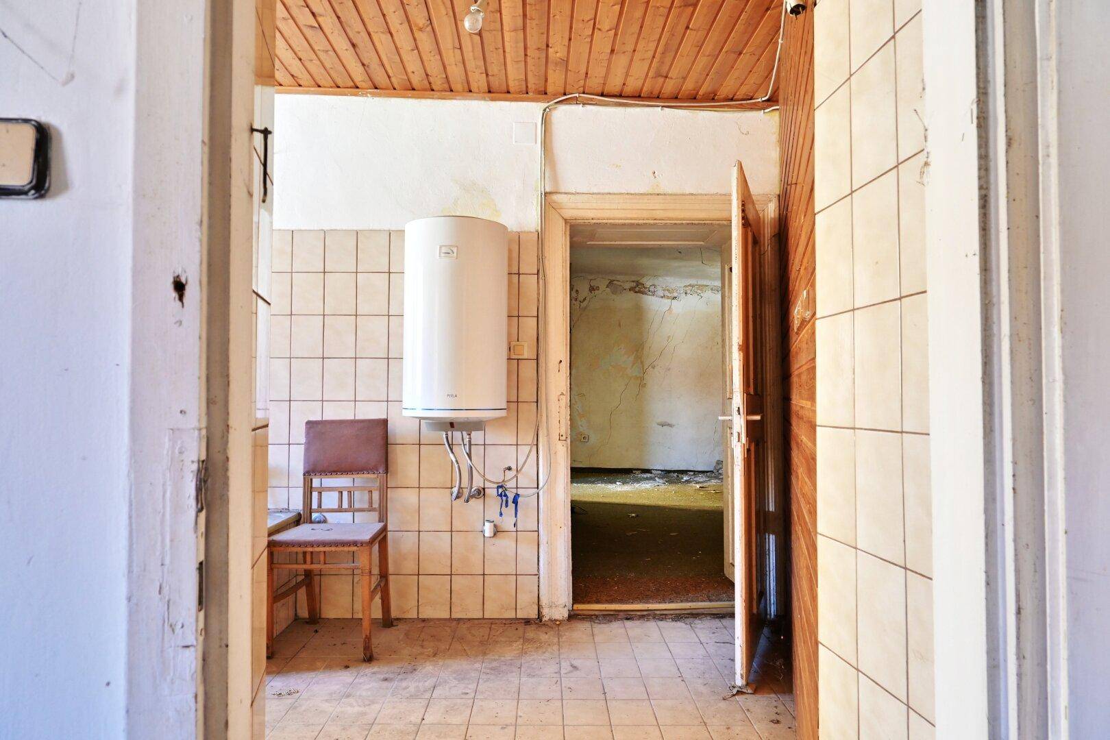 Hauswirtschaftsraum mit gefliesten Wänden, Warmwasserboiler und Holzdecke, der in einen weiteren Raum führt.