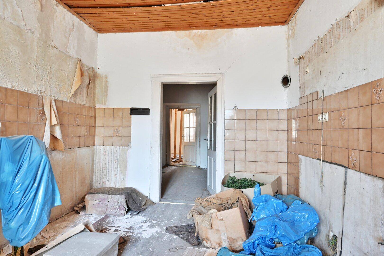 Renovierungsbedürftiger Raum mit gefliesten Wänden, abblätternder Tapete und Holzdecke, der auf eine Neugestaltung wartet.