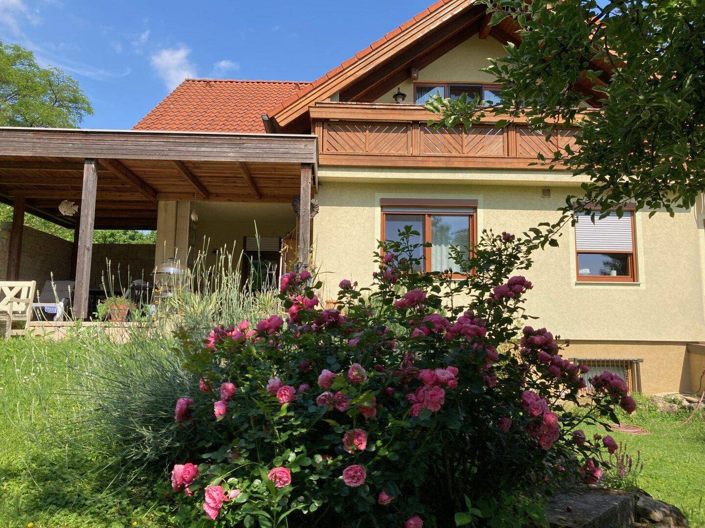 Hausfassade mit blühenden Rosenbüschen und überdachtem Terrassenbereich.