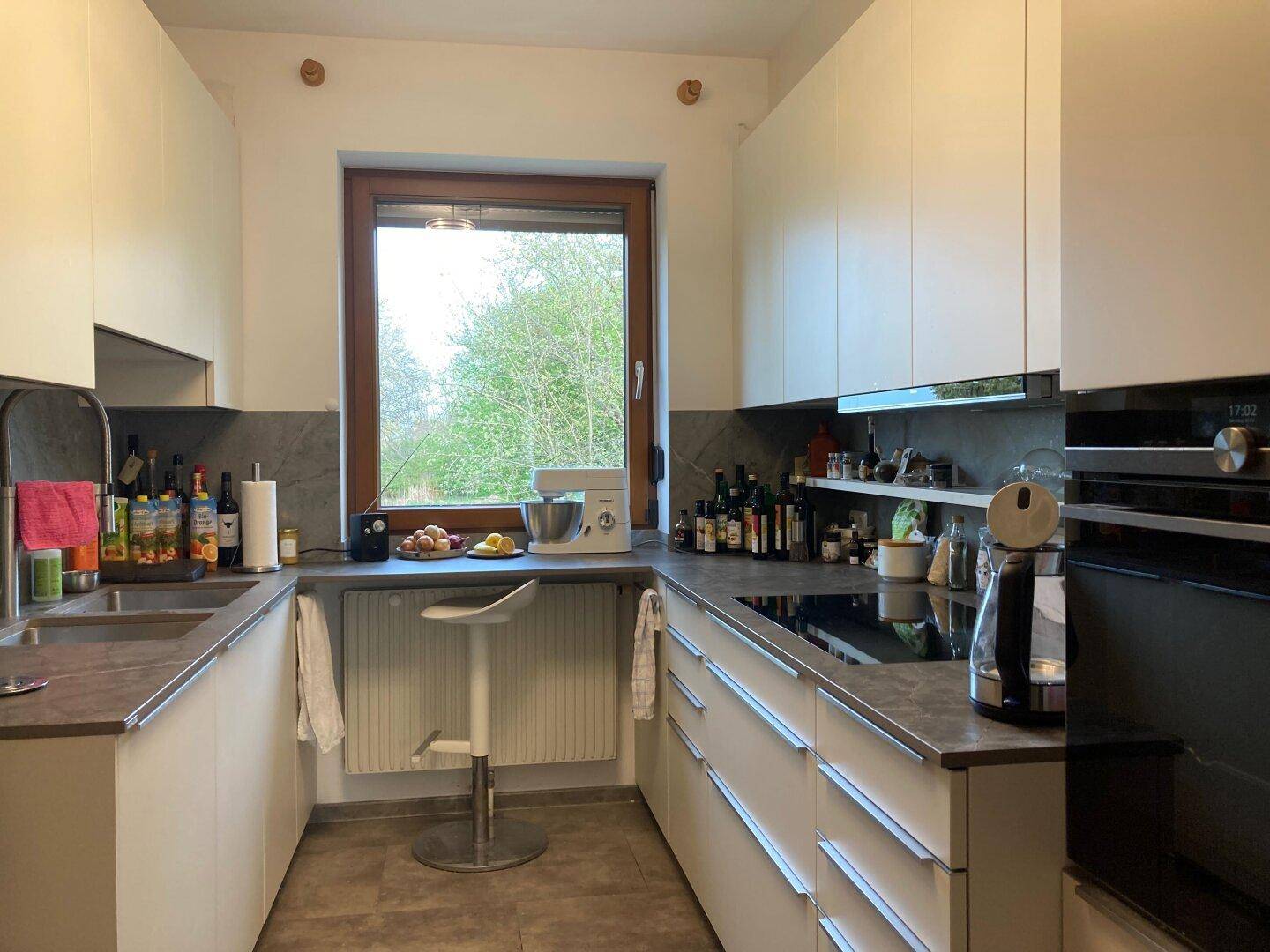 Moderne Küche mit weißen Schränken, Induktionskochfeld und Fenster mit Blick ins Grüne.