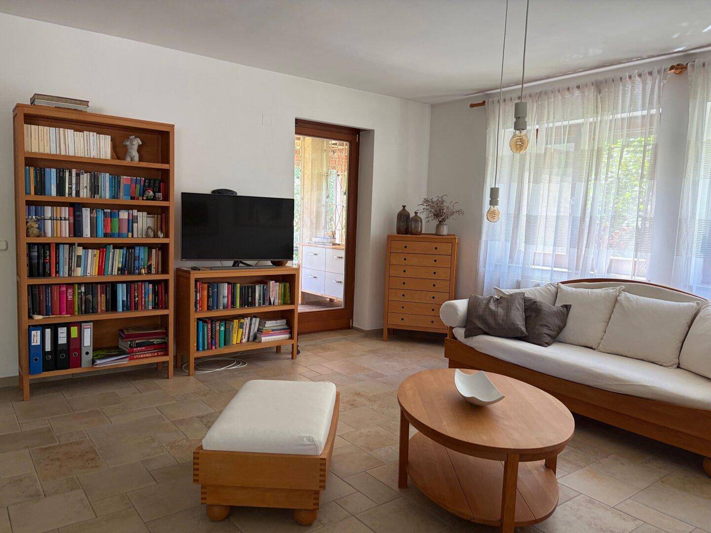 Heller Wohnbereich mit Sofa, Fernseher und Bücherregalen, offen zum Garten.