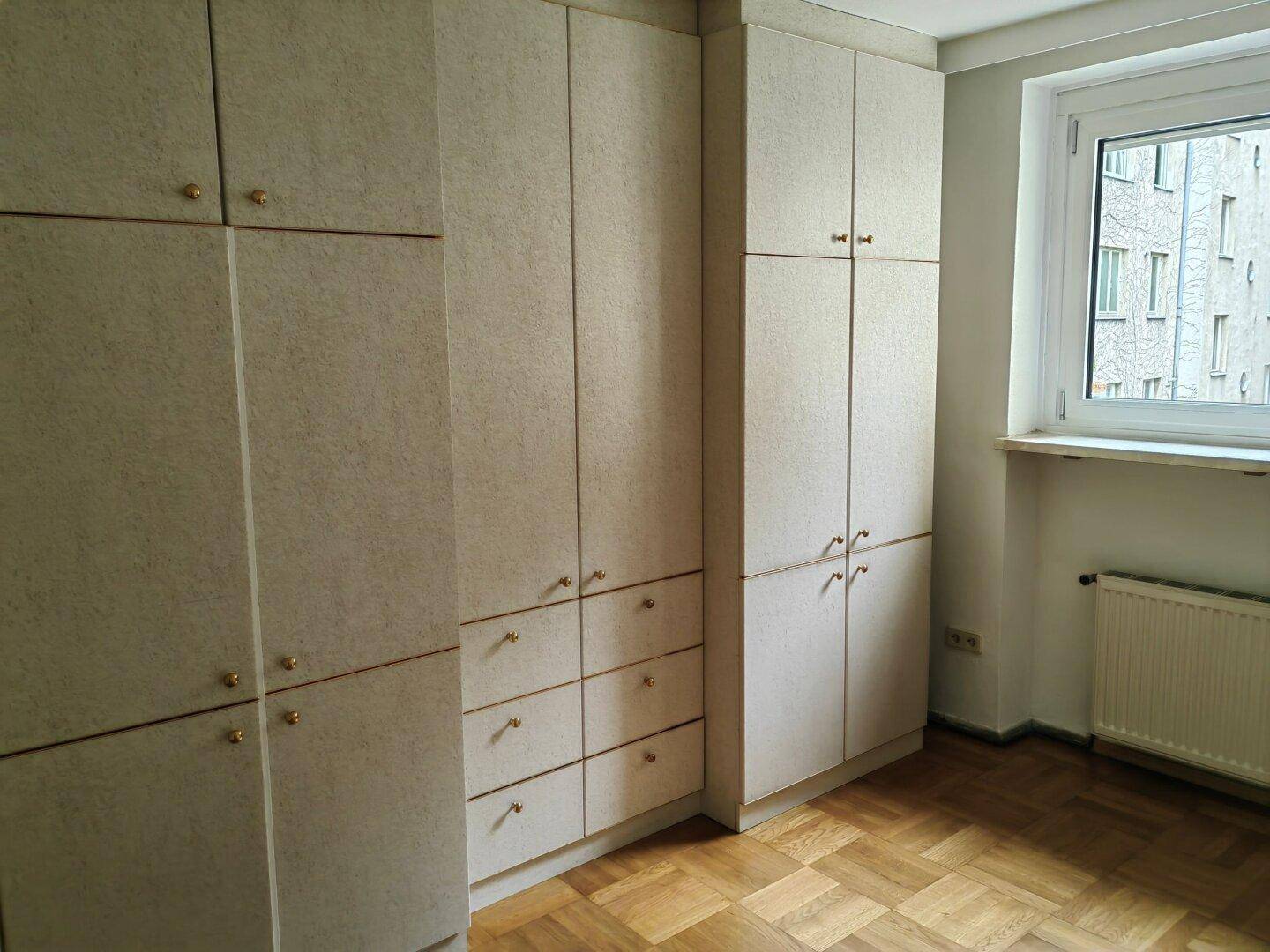 Schlafzimmer mit vielen hellen Einbauschränken, Parkettboden und einem Fenster für Tageslicht.