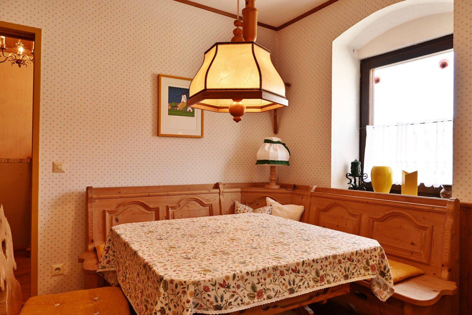 Detailansicht des Essbereichs mit traditioneller Holzeckbank, Tisch und floral gemusterter Tischdecke.