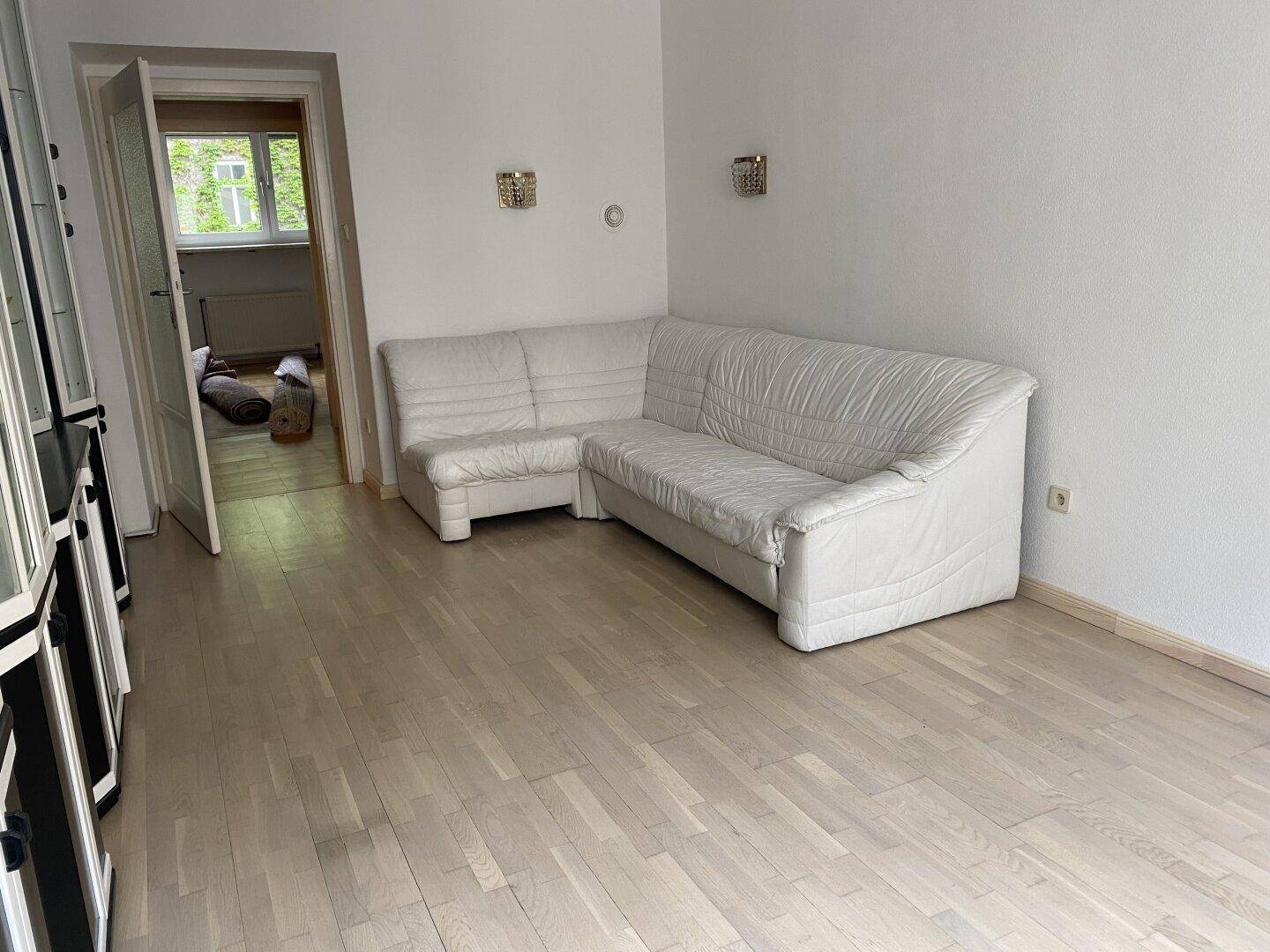 Heller Raum mit weißem Ecksofa und hellem Parkettboden, bietet Zugang zu einem weiteren Zimmer.
