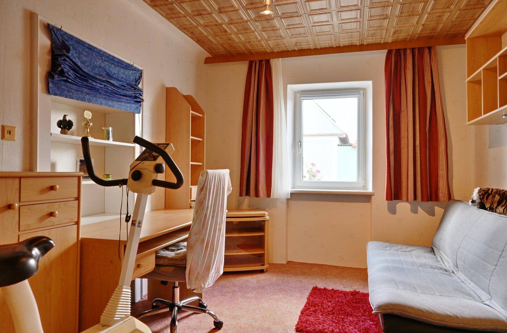 Schlafzimmer oder Arbeitszimmer mit Schreibtisch, Heimtrainer und Teppichboden.