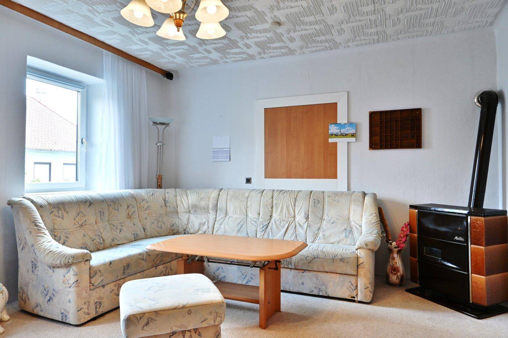 Helles Wohnzimmer mit Ecksofa, Kachelofen und traditioneller Deckenverkleidung.