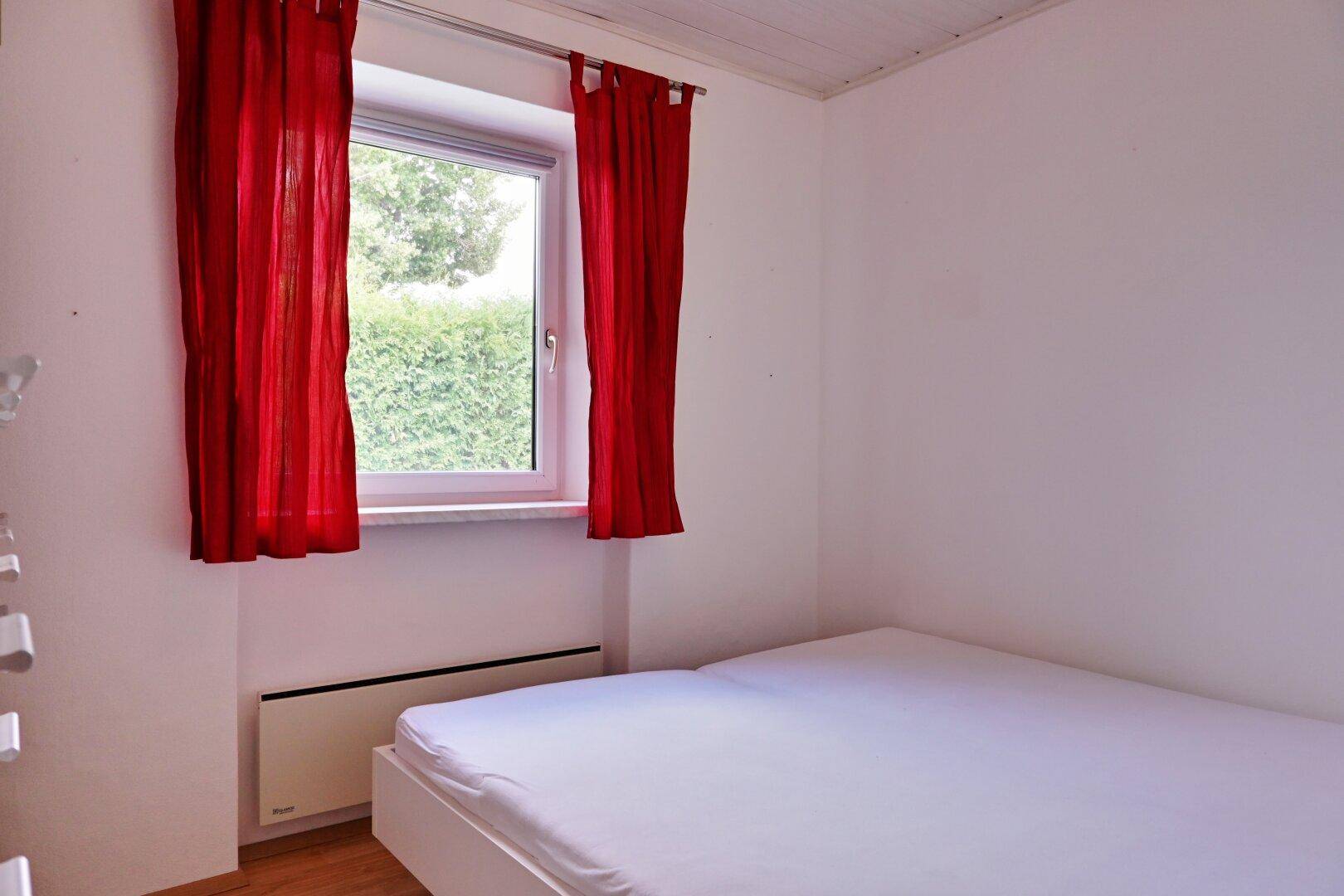 Einfaches Schlafzimmer mit weißem Bett, Holzfußboden und Blick ins Grüne.