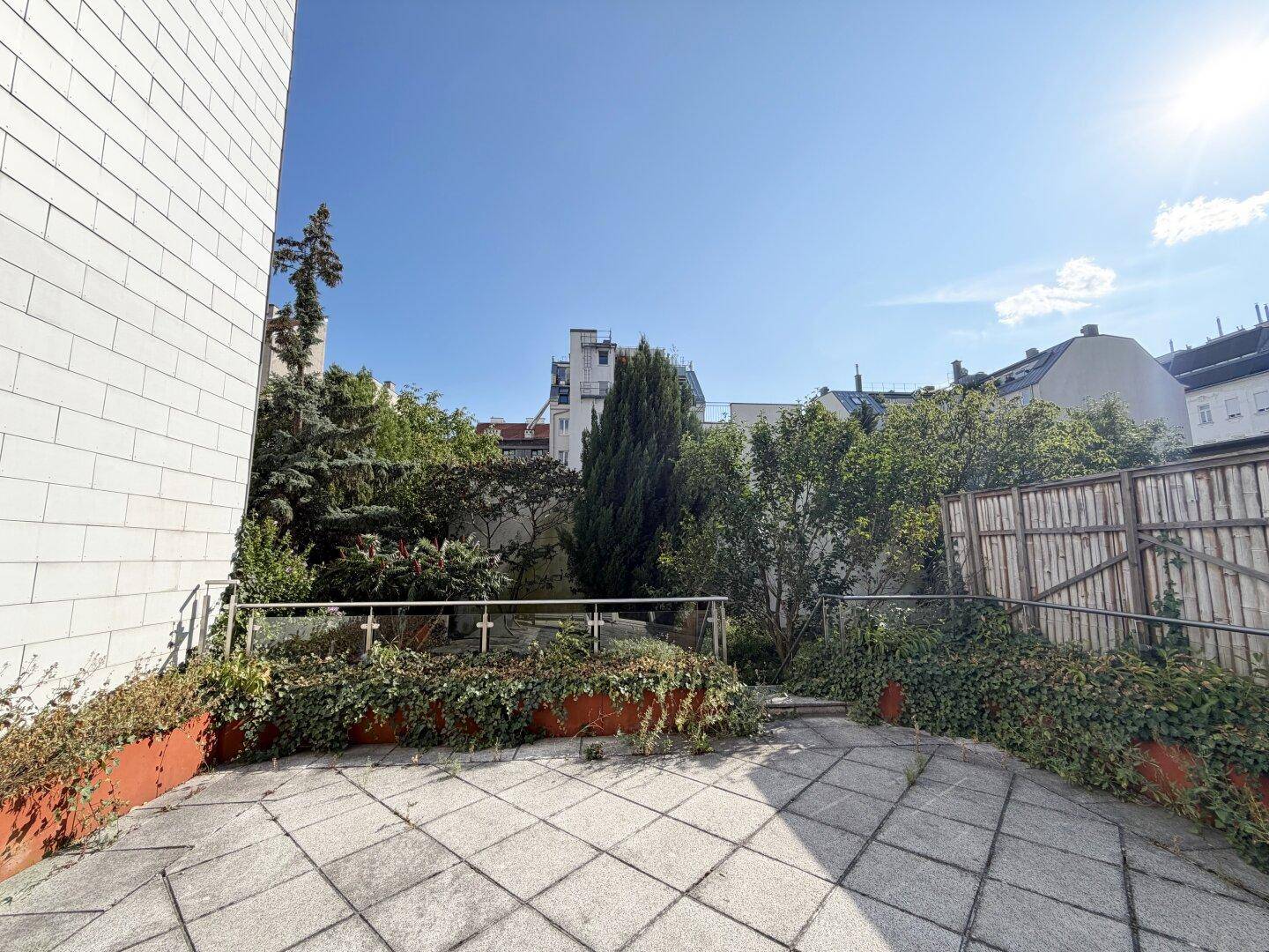 Sonniger Außenbereich mit gepflasterter Terrasse und üppiger Bepflanzung, umgeben von Gebäuden.