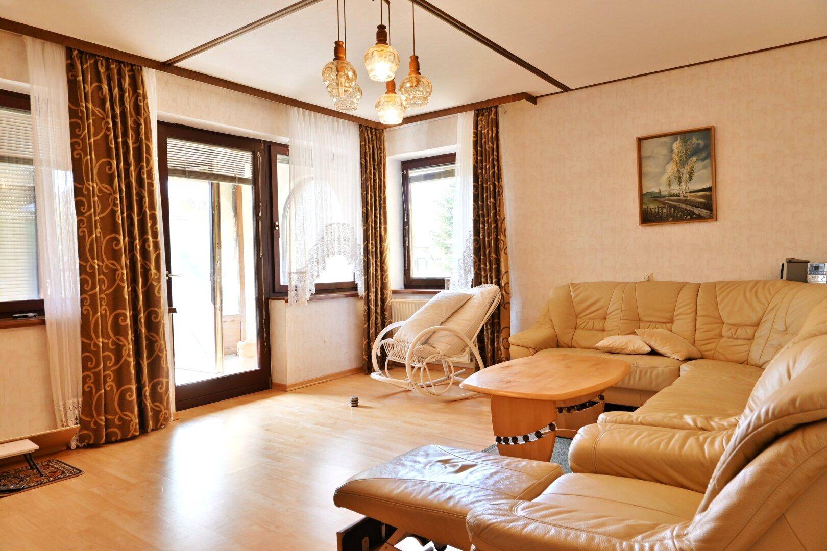 Gemütliches Wohnzimmer mit heller Ledercouch, Parkettboden und Zugang zu einem Balkon.
