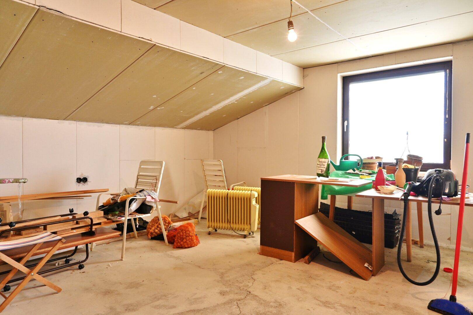 Dachbodenraum mit schräger Decke und Fenster, genutzt als Abstellfläche.