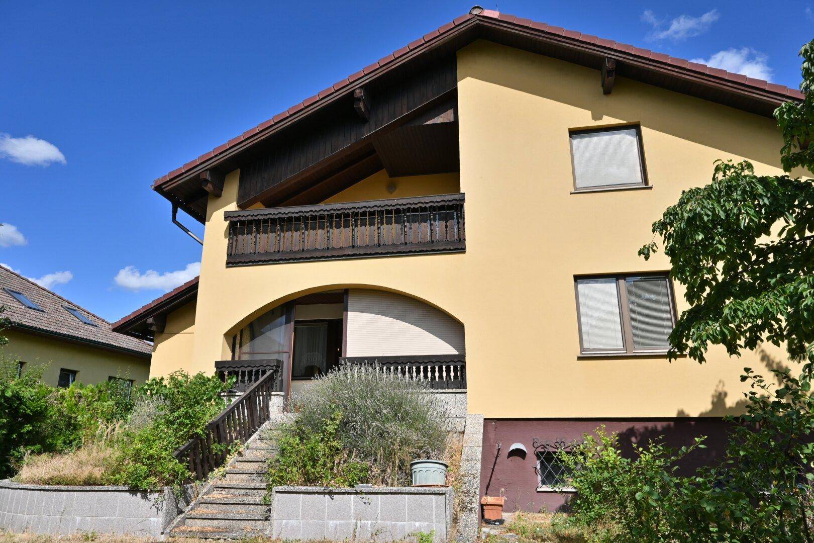Gelbe Hausfassade mit traditionellem Holzbalkon und einem überdachten Eingangsbereich im Erdgeschoss.