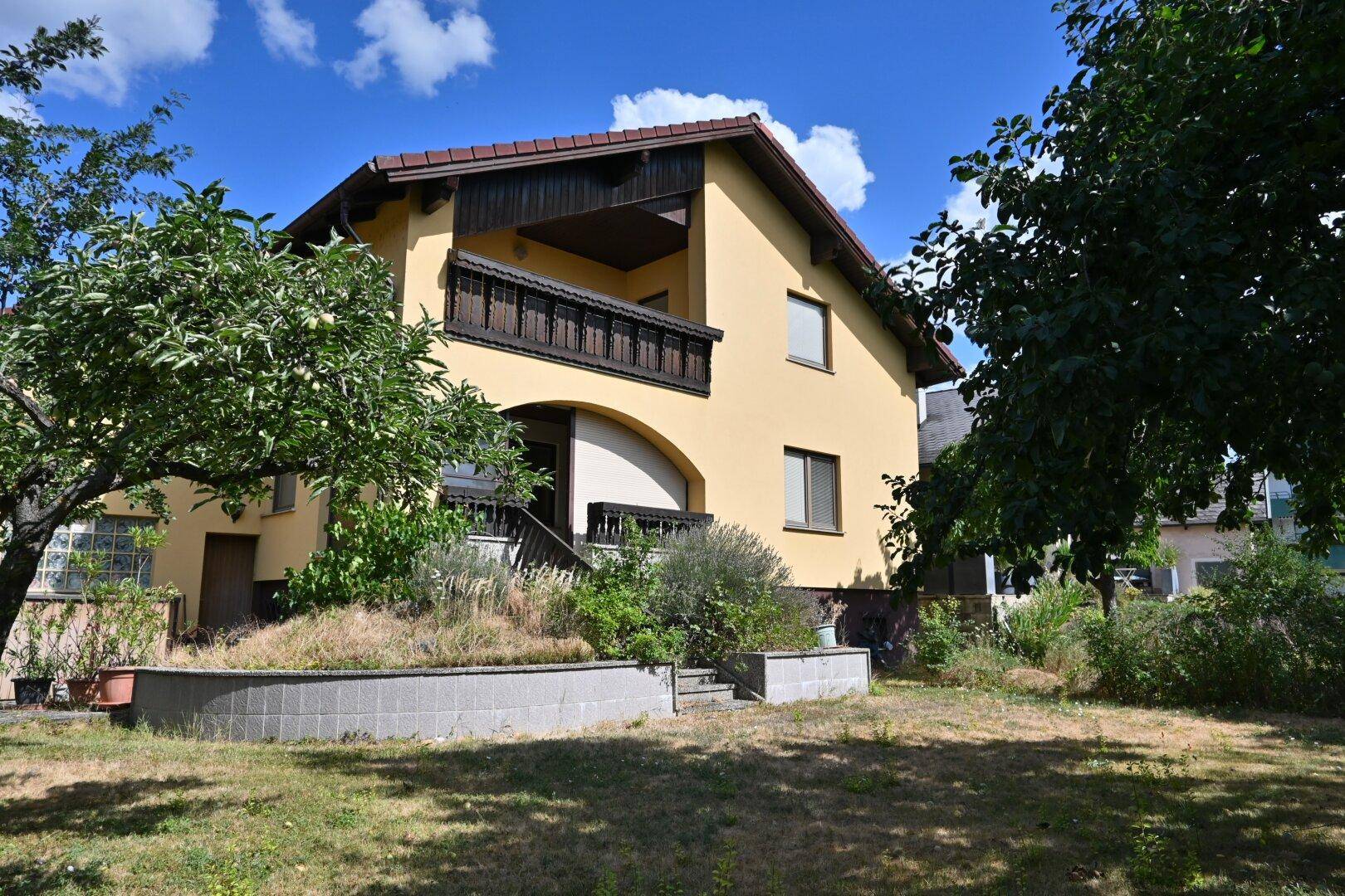 Ansicht des gelben Hauses mit Balkon und großem Gartenbereich mit Bäumen und Sträuchern.