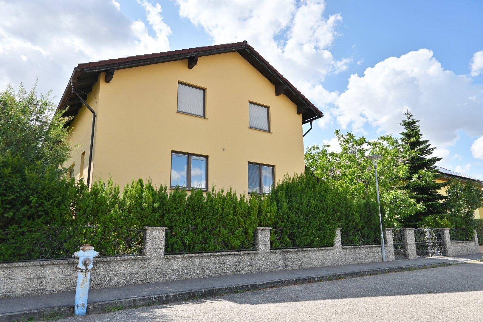 Gelbes Haus von der Straße aus gesehen, umgeben von einer grünen Hecke unter blauem Himmel.