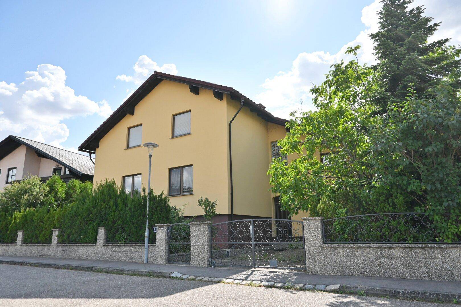 Hausansicht von der Straße mit gelber Fassade, schmiedeeisernem Tor und grüner Bepflanzung.