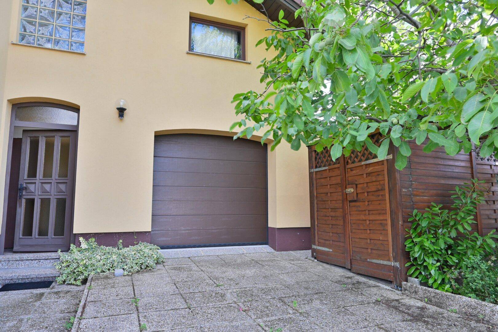 Garagentor und Hauseingang mit gepflastertem Vorplatz und einem Baum im Vordergrund.