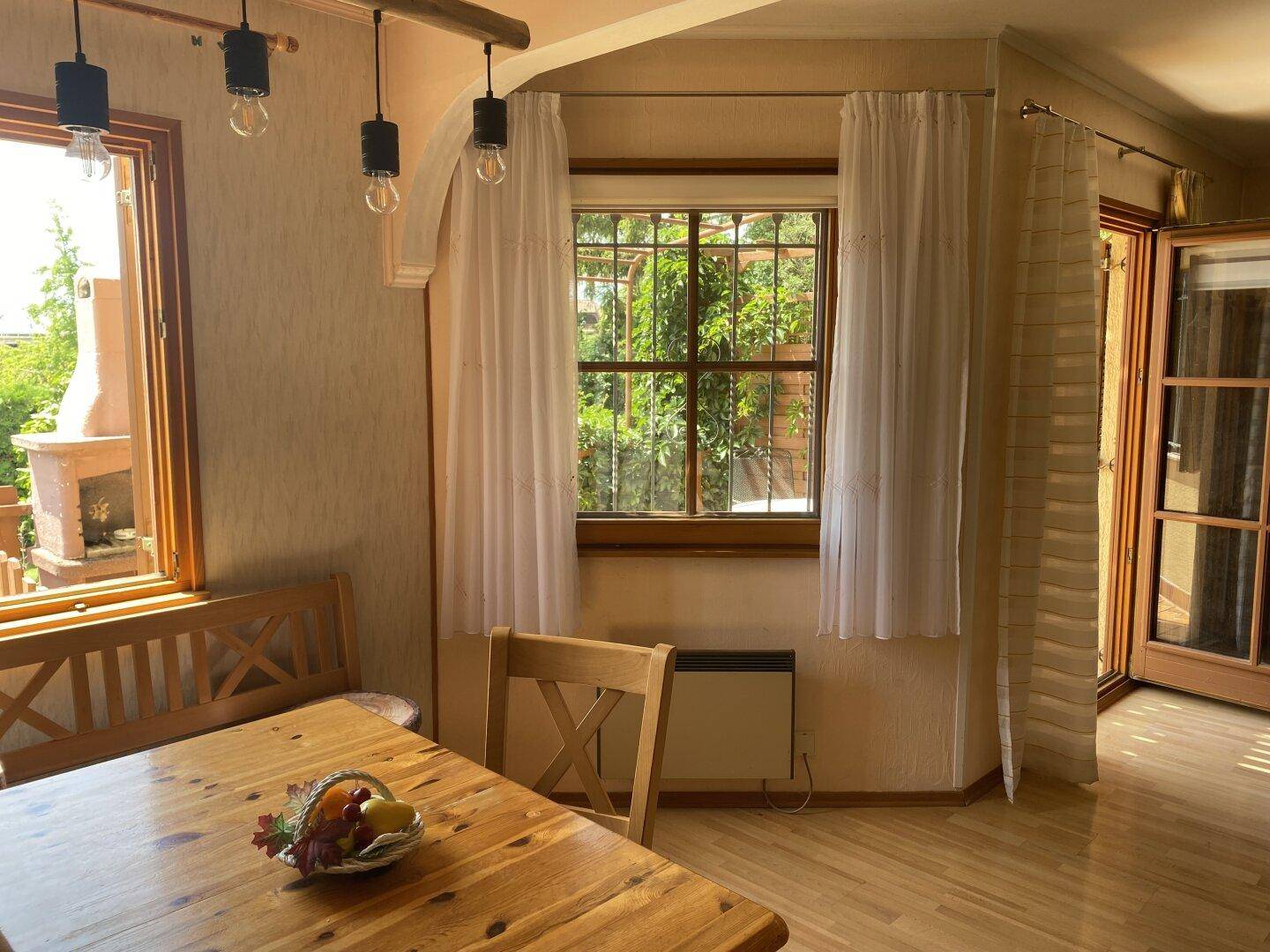 Heller Essbereich mit Holztisch und Bänken, umgeben von Fenstern mit Vorhängen.