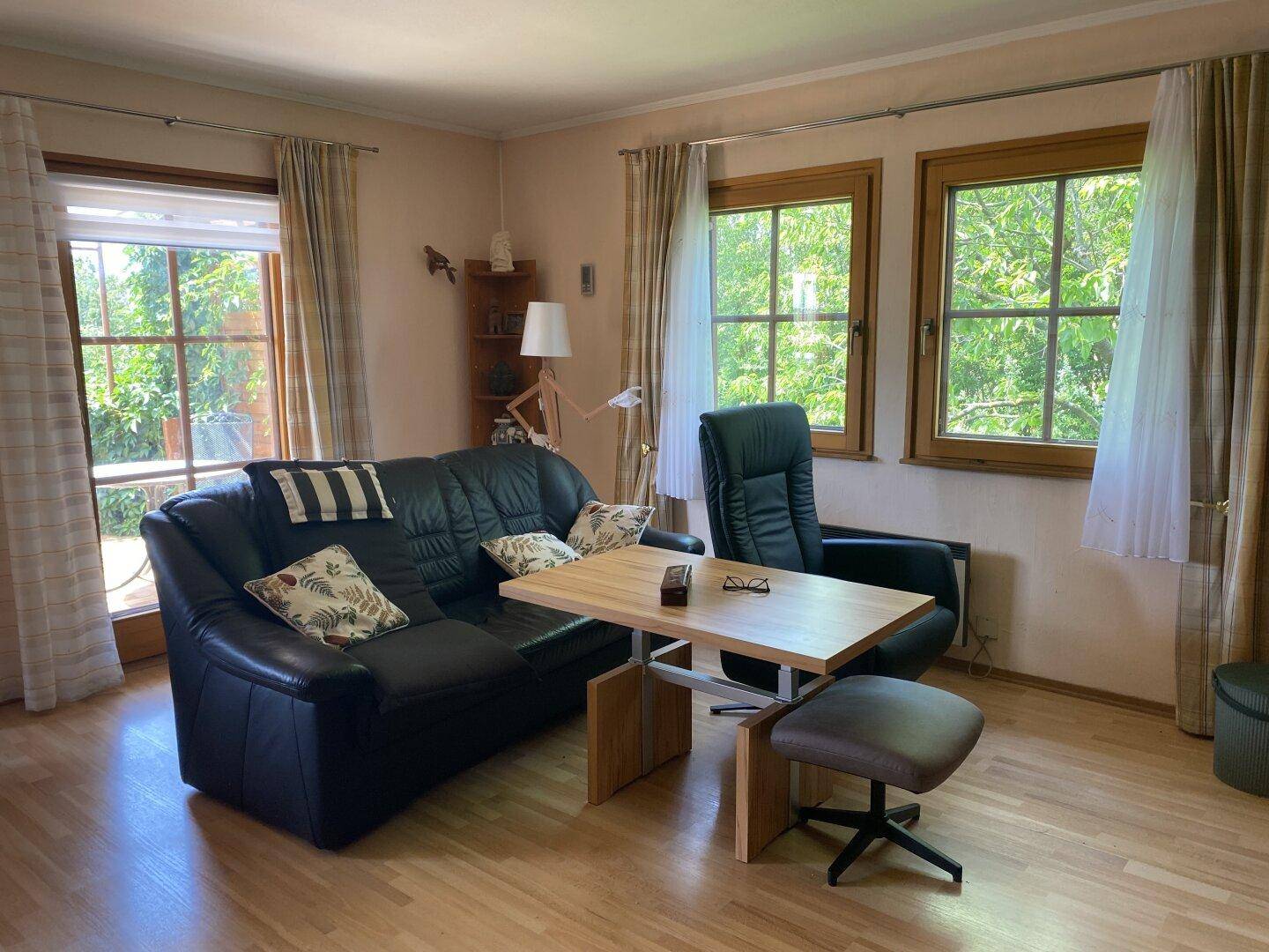 Helles Wohnzimmer mit Ledersofa, Holzboden und Fenstern mit Blick ins Grüne.