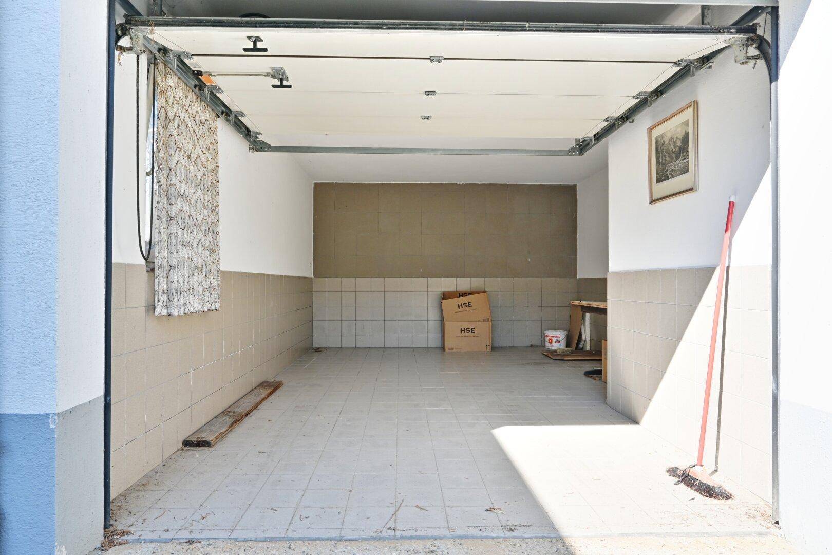 Leere Garage mit aufgerolltem Tor, Fliesenboden und einfacher Ausstattung.